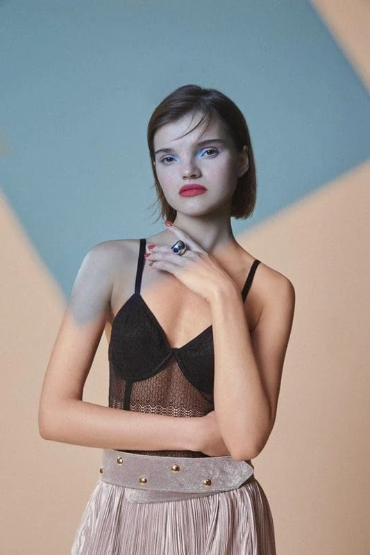 A young woman with short brown hair, wearing a black lace top and beige pleated skirt, posing confidently for a fashion magazine.