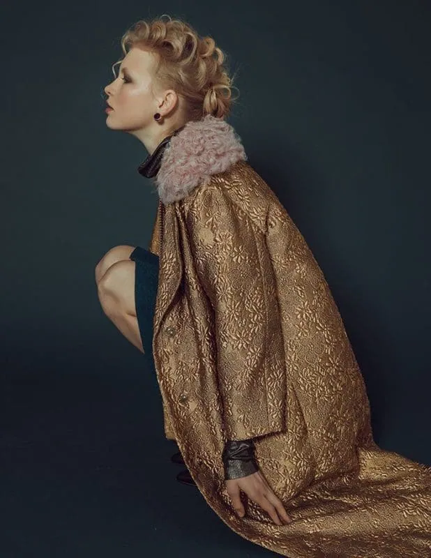 Fashion model wearing ornate gold coat with pink fur collar, stylishly posed for Vogue.