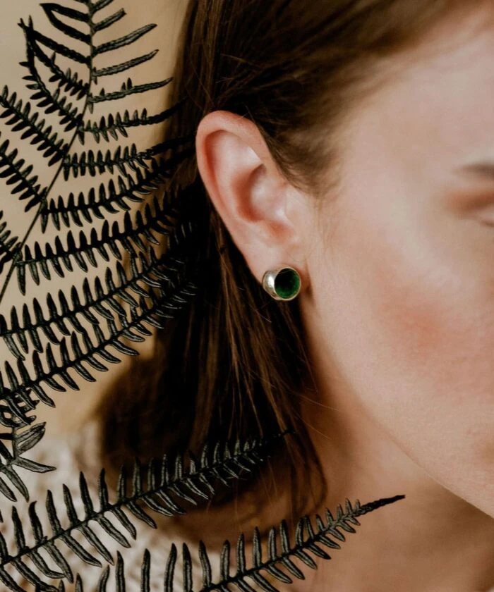 Close-up of a woman wearing small green stud earrings with fern leaves in the background.