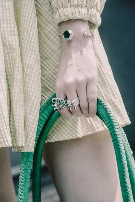 Close-up of a woman's hand holding a green gardening hose, stylish jewelry, and a yellow checkered jacket.