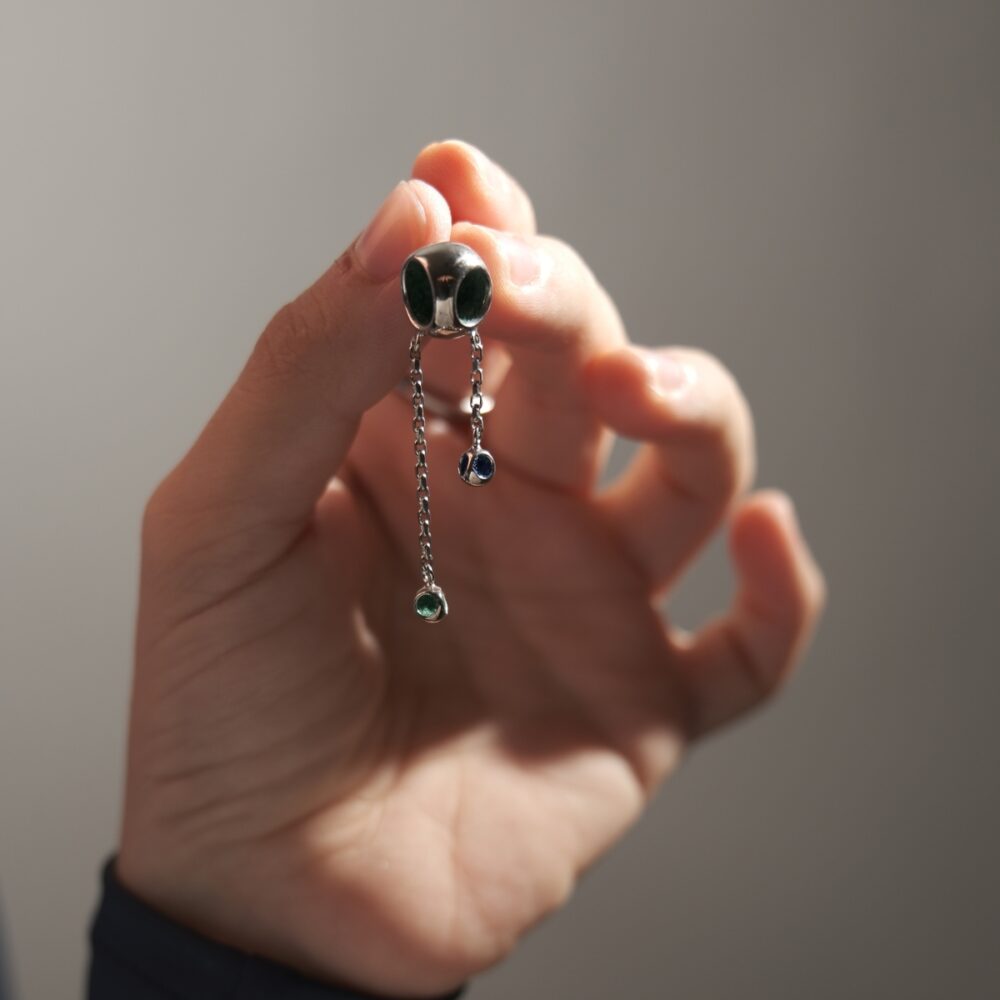 Close-up of modern silver chain earrings featuring colorful gemstone drops, held by a hand against a neutral background.
