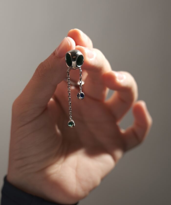 Close-up of modern silver chain earrings featuring colorful gemstone drops, held by a hand against a neutral background.