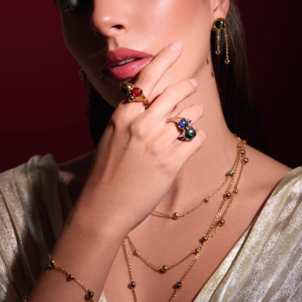 Close-up of a woman wearing luxurious gold and gem jewelry including rings, necklaces, and earrings with colorful stones.
