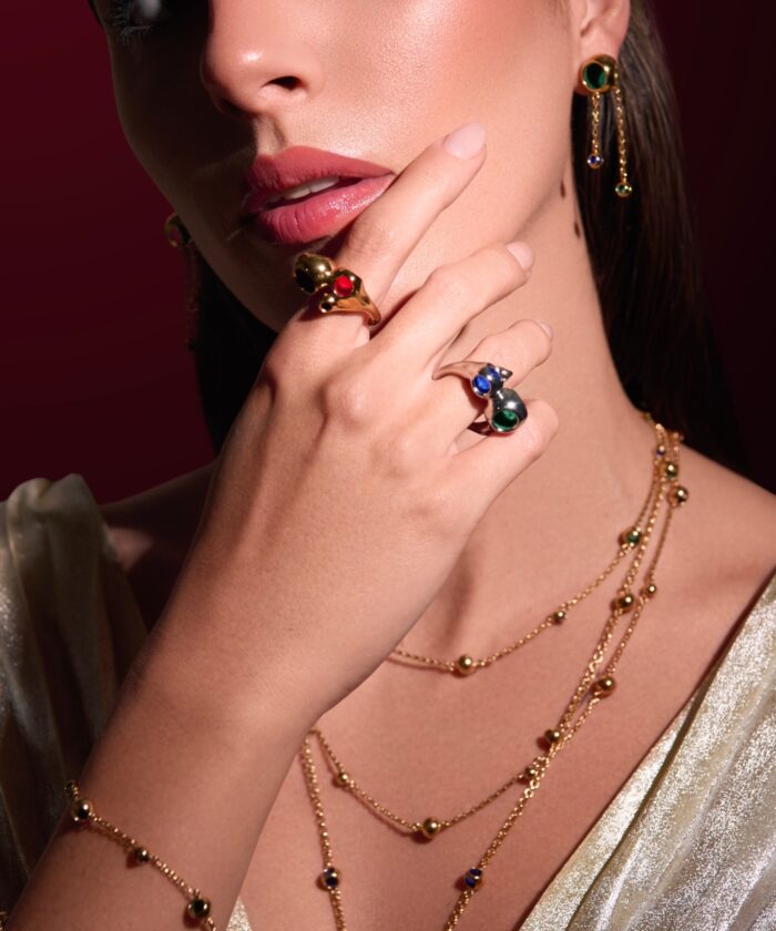 Close-up of a woman wearing luxurious gold and gem jewelry including rings, necklaces, and earrings with colorful stones.