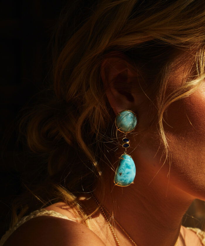 Dazzling Larimar earrings on Monica Varela wearing gold earrings with blue stones, showcasing vibrant jewelry.