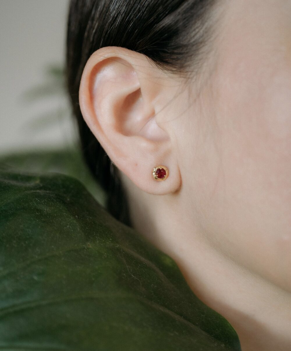 Close-up of Monica Varela ruby stud earrings featuring red gemstones in a gold setting, worn by a woman.