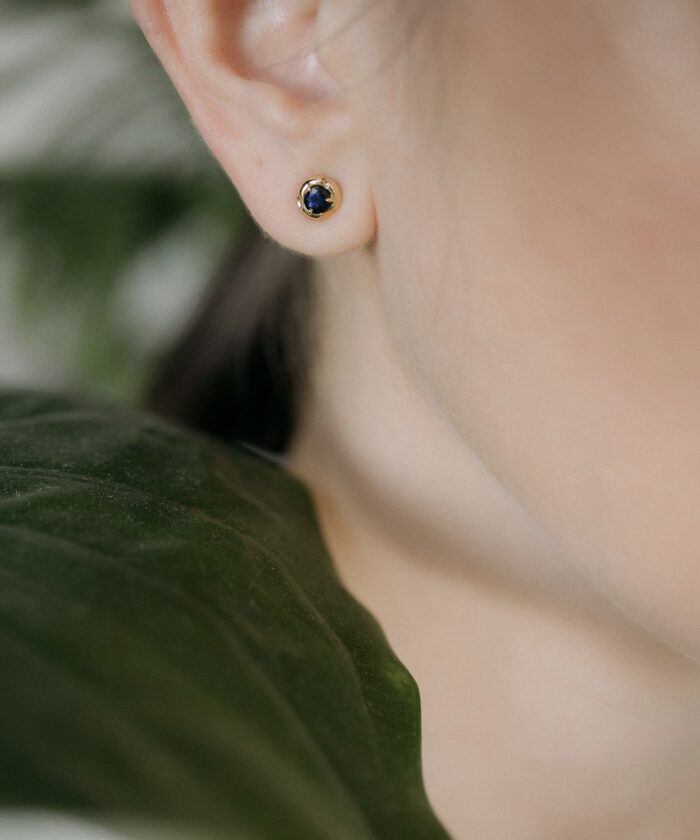 Delicate gold and sapphire stud earrings on a woman's earlobe, showcasing luxury jewelry.