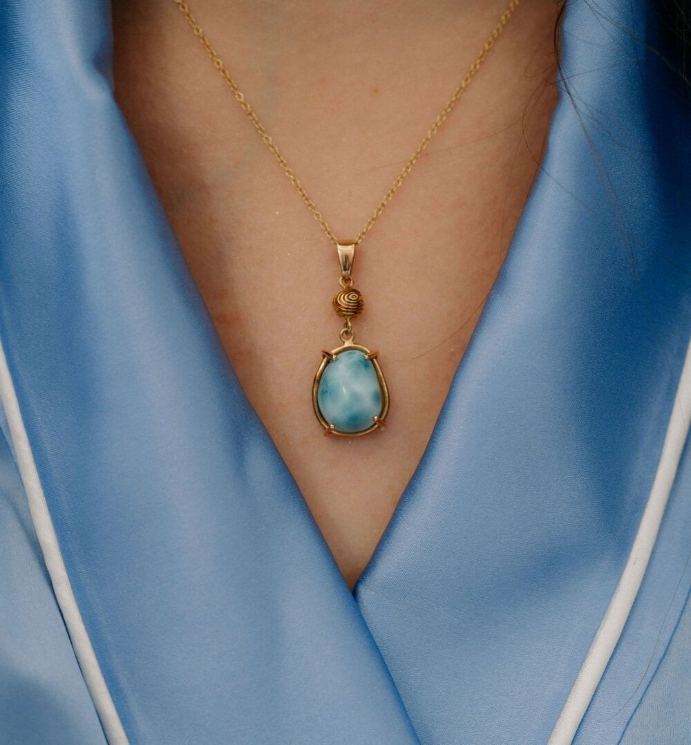 Close-up of a gold pendant necklace featuring a stunning larimar gemstone, worn against a silky blue blouse.