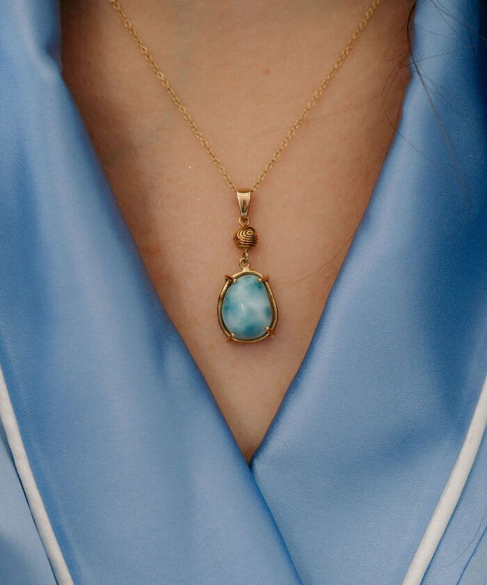 Close-up of a gold pendant necklace featuring a stunning larimar gemstone, worn against a silky blue blouse.