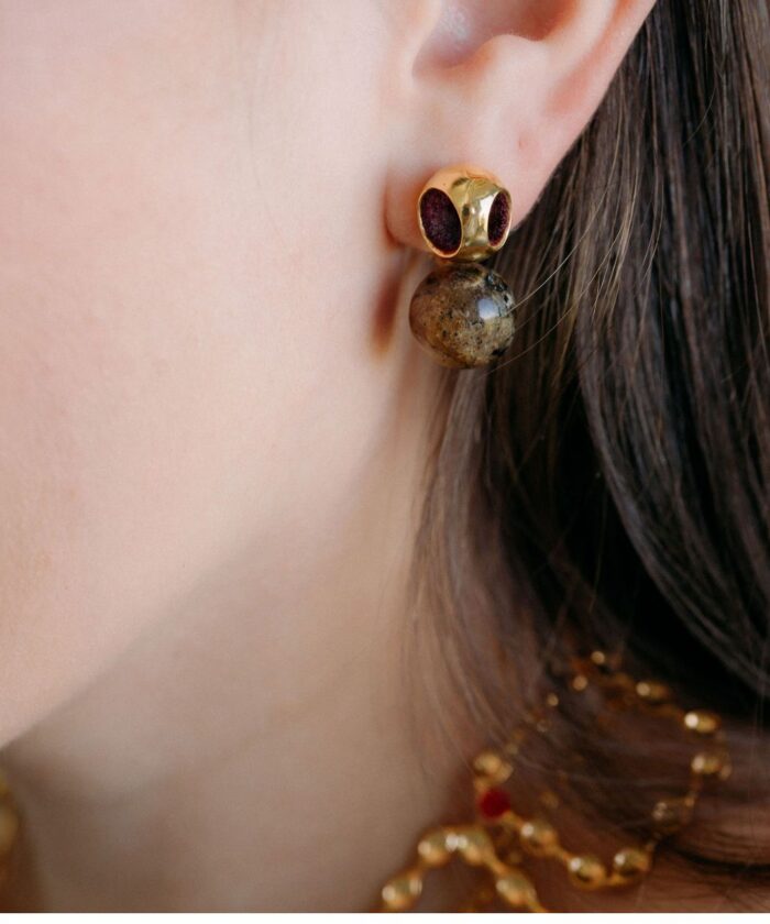 A close-up of a woman's ear with gold and gemstone stud earrings, showcasing luxury jewelry with dark red and brown details.