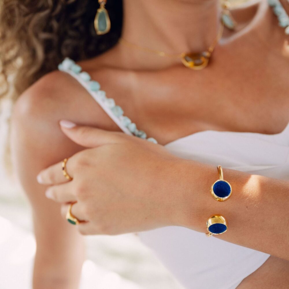 Close-up of gold bracelet and rings featuring blue stone details, worn with a white dress and gold necklace.