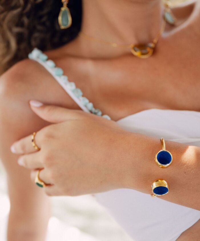 Close-up of gold bracelet and rings featuring blue stone details, worn with a white dress and gold necklace.