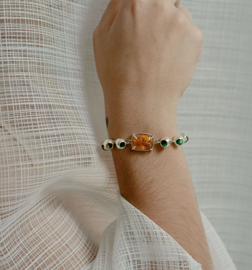 A close-up of Monica Varela wearing a stunning amber and silver infinity bracelet with green accents, styled on a delicate person’s wrist.