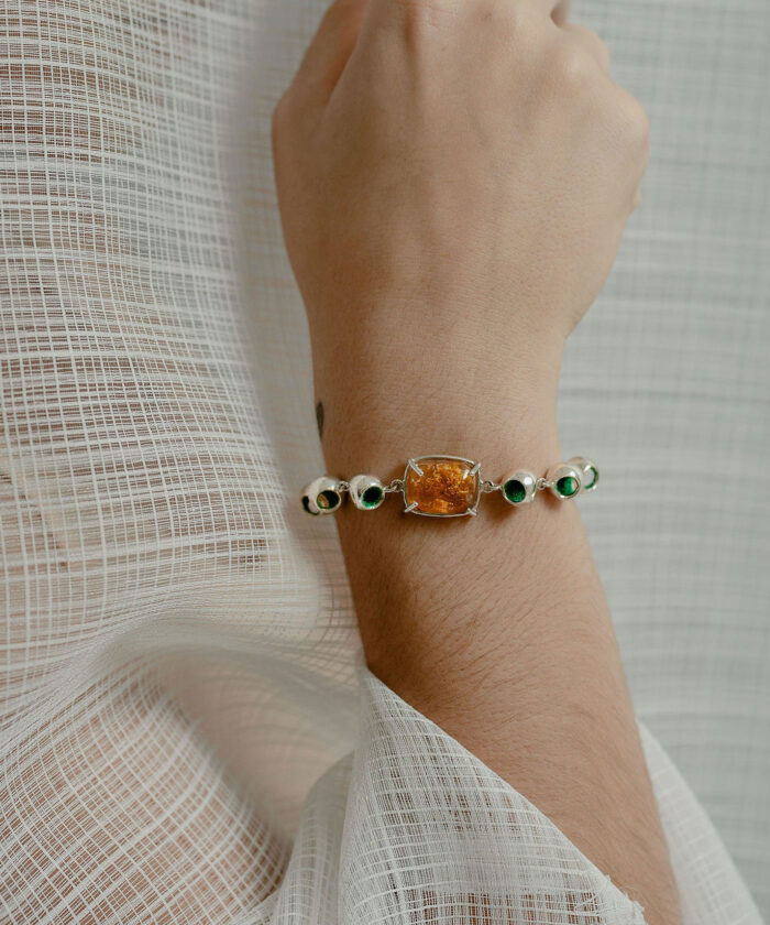 A close-up of Monica Varela wearing a stunning amber and silver infinity bracelet with green accents, styled on a delicate person’s wrist.