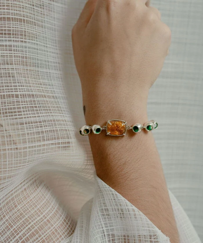 A close-up of Monica Varela wearing a stunning amber and silver infinity bracelet with green accents, styled on a delicate person’s wrist.