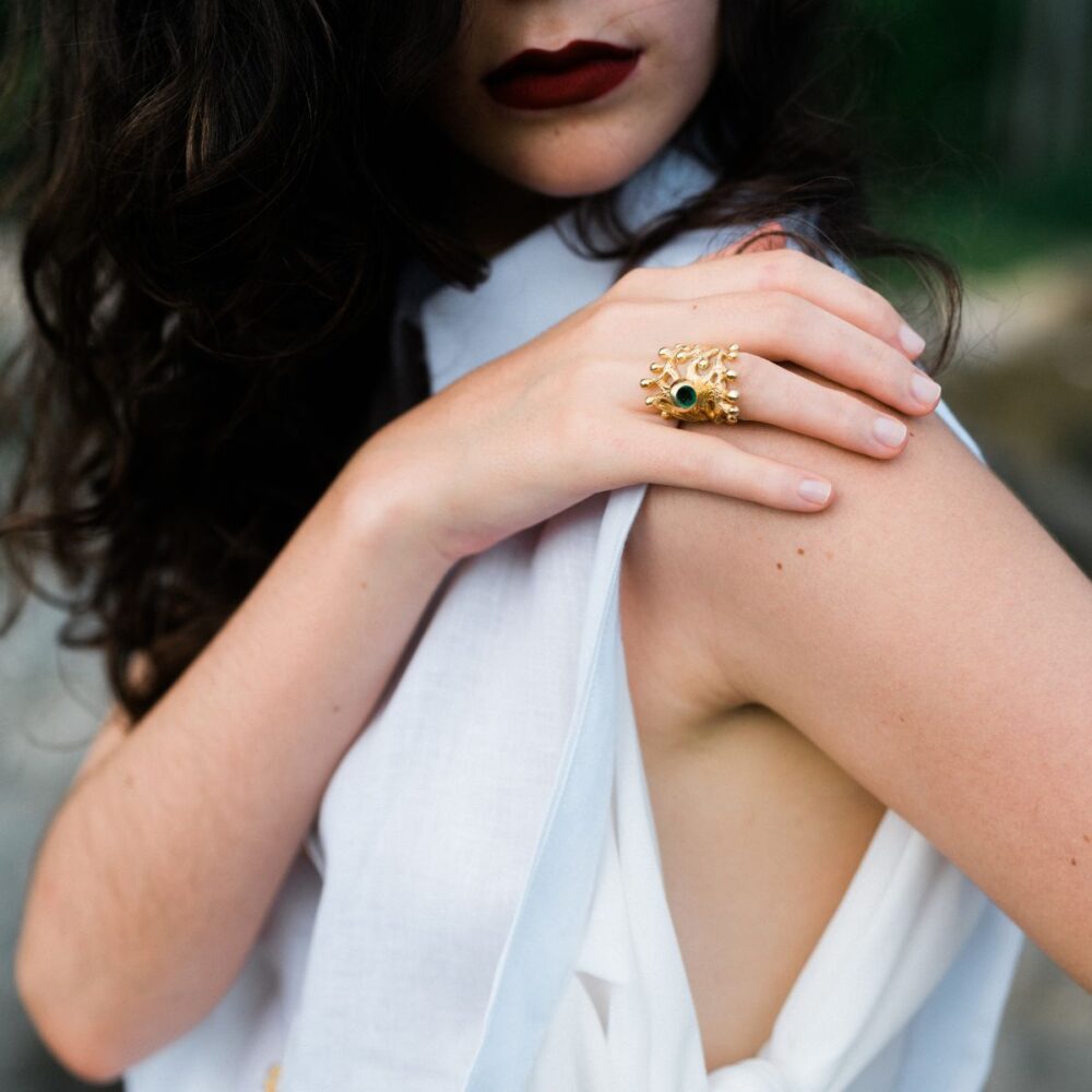 Close-up of woman wearing bold gold snake ring with green gemstone, emphasizing jewelry details.