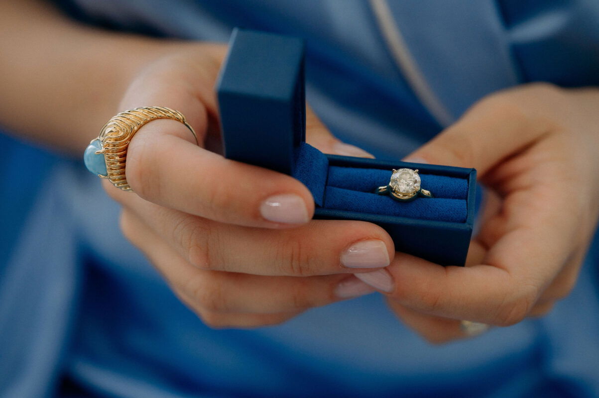Close-up of a woman's hand holding a blue jewelry box with a sparkling diamond ring inside.