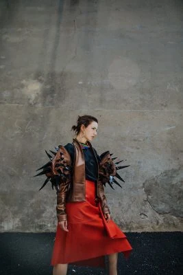 Monica Varela wearing bold fashion and accessories, striking pose against urban concrete wall.
