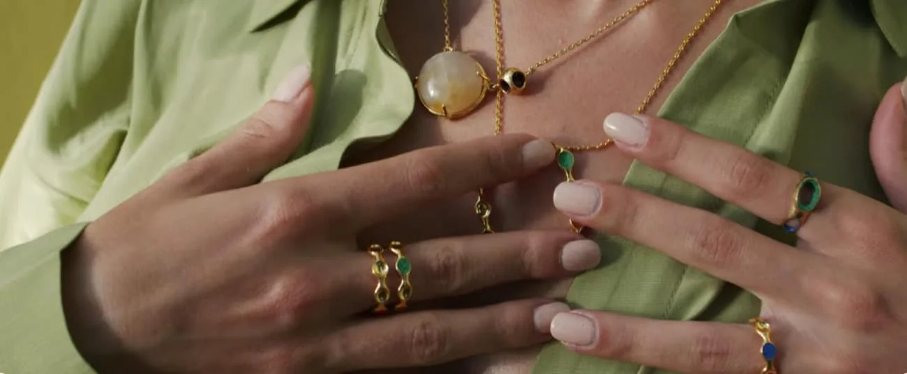 Close-up of gold rings and necklaces with colorful gemstones on elegant woman's hands and neckline.